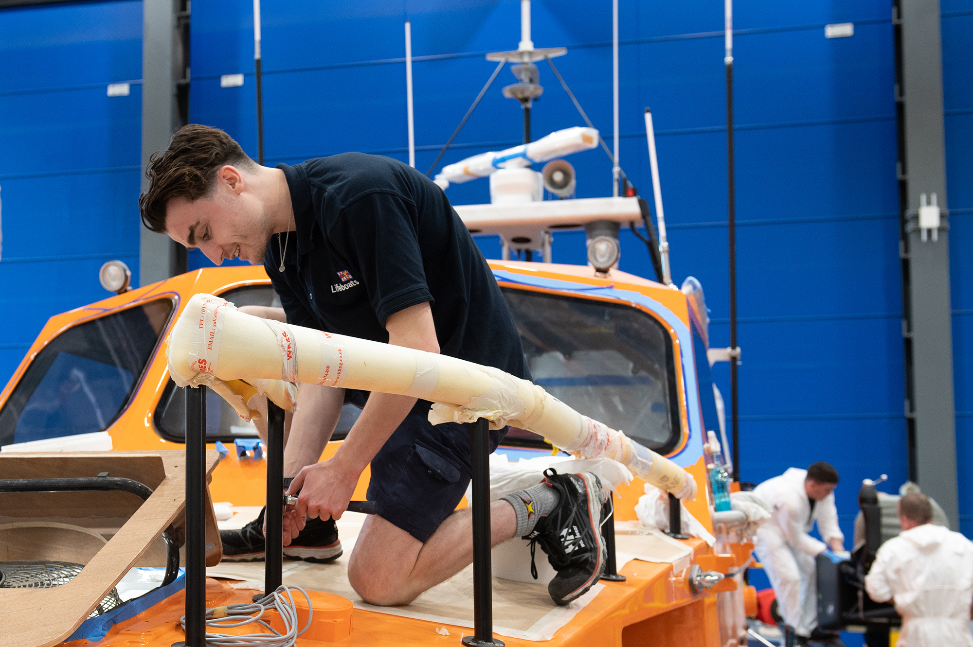 An apprentice kneels on a lifeboat deck working. The boat is still under construction. In the background 2 people in white overalls work on an engine