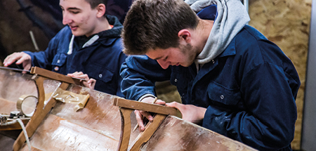 Students in overalls fix a section of a wooden boat