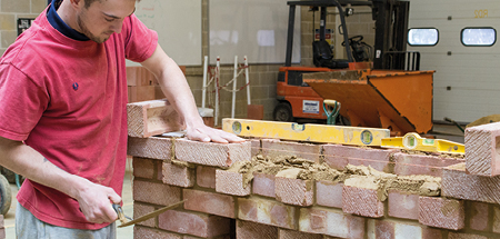 Student holding a trowel presses a red brick on to the top of a brick wall he is constructing