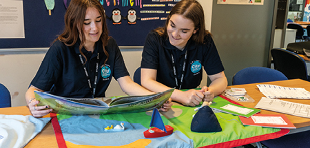 2 students work with a story sack. They are reading a book, with fabric mat depicting a water and land scene, a toy boat and swans and flash cards spread on the table infront of them