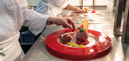 Students add garnish to a decorative plate containing a desert in a restaurant kitchen