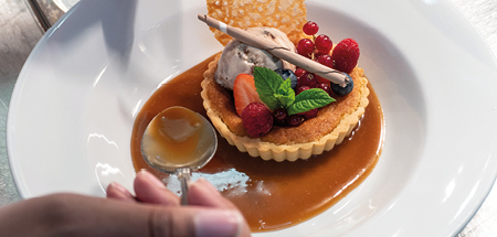 Sauce is poured from a spoon around the edge of a beautiful tart with berries, ice cream, brandy snap and mint on top