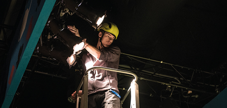 Student stands on a tower wearing a hard hat and harness, and positions stage lights