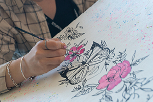 Close-up of a hand with a paint brush. Working on a canvas with outlines drawn of flowers and a butterfly obscuring the top half of a face, painting one of the flowers pink