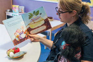 Student sits at a table reading an illustrated story book to a life-size toddler doll that sits on her lap
