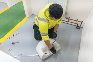 Student in high-vis and gloves uses an adjustable spanner on copper pipes to attach them to a hand basin that is upside down on the floor