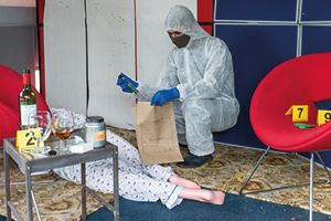 Student wearing a white, hooded, paper crime scene overall, mask and gloves places an object into a brown paper evidence bag. There is a body laying on the floor and a table with wine bottle and glasses and yellow cards with crime scene number