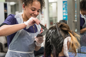 Student wearing a plastic apron and gloves uses a paddle and brush to colour a client’s hair