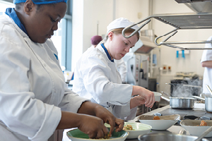 Students in chef’s whites plate up food. In the background you can see saucepans with steam rising from them