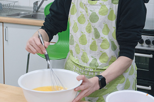 Student in an apron whisks eggs in a bowl. In the background is a domestic cooker