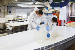 2 students in white protective suits and gloves lay a sheet of material inside a white boat hull