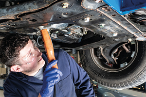 Student with protective eye wear, gloves and overalls. He stands underneath a car, in a workshop, using a torch to inspect the underside
