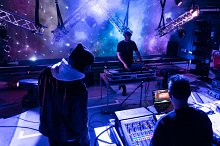 Studio space set up with a stage and architectural design and creative blue and purple lighting. A student operates the lighting desk with a DJ on decks in front of the stage