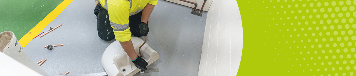 Student in high-vis and gloves uses an adjustable spanner on copper pipes to attach them to a hand basin that is upside down on the floor