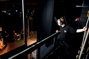 A student wearing a headset looks down from an overhead gantry onto the stage