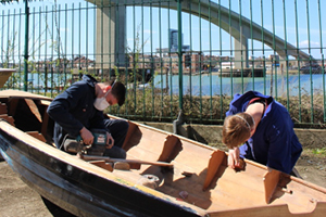 2 students sand down a wooden boat outdoors
