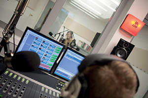 A student at a mixing desk looking through to another student inside the audio recording studio, a lit up sign on the wall reads Mic Live