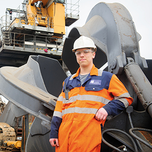 An apprentice wearing a hard hat and overalls stands in front of large plant machinery